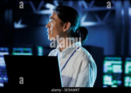 Cheerful BIPOC man between server hub rows providing processing resources for businesses worldwide. Computer scientist fixing data center mainframes tasked with managing massive databases Stock Photo