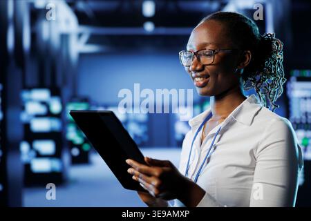 Smiling woman between server hub rows providing processing resources for businesses worldwide. Happy admin fixing data center mainframes tasked with managing massive databases Stock Photo