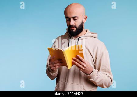 Young thoughtful focused arab man reading novel from book with yellow cover. Concentrated bald bearded person holding open softcover textbook and studying with pensive expression Stock Photo