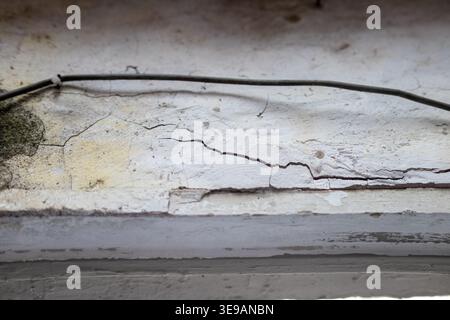 Detailed view of deep cracks and peeling paint on a weathered white concrete surface with a thin black cable, representing structural issues, house ma Stock Photo