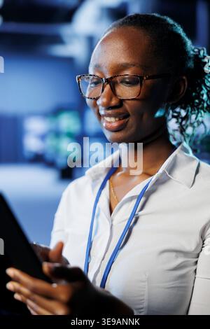 Pleased admin in data center uses tablet to prevent system hardware overload. Seasoned expert between server supercomputers ensuring enough network bandwidth for smooth operations Stock Photo