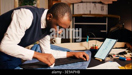 Male couturier marking fabric design to craft fashion items in tailoring atelier, using chalk to measure garment for needlework. Designer working on clothing line creation, workshop. Stock Photo