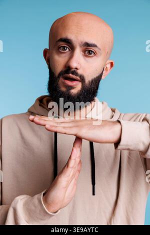 Arabian person taking time out with stop signal while showing confident facial expression portrait. Young bald bearded man making interruption gesture with hands while looking at camera Stock Photo
