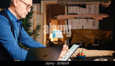 Senior tailor designing sketches of clothes using tablet and laptop, working on modern fashion collection in luxury atelier. Professional designer creating clothes with tailoring tools. Handheld shot. Stock Photo