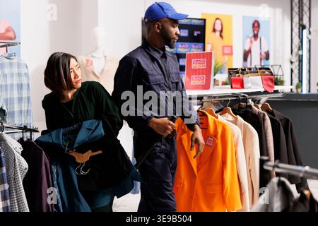 Anxious Asian woman shopper holding clothes in hands looking nervously aside, standing near security guard in mall. Female customer feeling stressed while shopping in clothing store on Black Friday Stock Photo