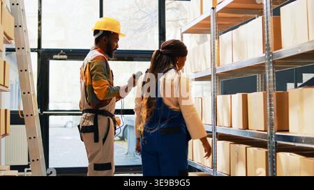 Team of employees scanning merchandise in depot, using scanner and tablet to check barcodes for stock logistics. Man and woman working on inventory, cargo pallets on racks and shelves. Handheld shot. Stock Photo