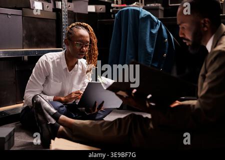 Detective and assistant sitting on floor surrounded by case files and working overtime in agency office. Two african american cops making research, searching crime solving solution Stock Photo