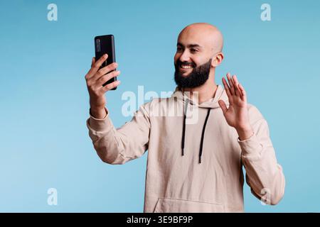 Smiling arab man waving hi while talking online using smartphone videocall application. Friendly person having remote communication, greeting with hand and speaking with mobile phone front camera Stock Photo