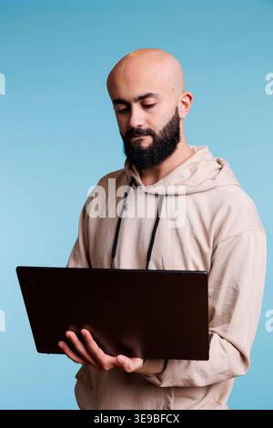 Concentrated arab man reading business report on laptop and typing online message. Focused young person holding portable computer, using software apps and browsing internet Stock Photo