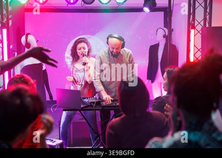 Woman singing and man dj mixing tracks while people partying on dancefloor in nightclub. People dancing and enjoying electronic music band show on stage with spotlights in club Stock Photo