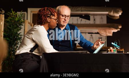 Craftsman teaching trainee to sew using textile industry tools and create fashion design. Beginner learning to manufacture clothing with experienced tailor in workshop. Handheld shot. Stock Photo