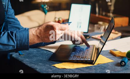 Male tailor buying fabric on website with credit card at laptop, doing shopping for material and textile industry tools for luxury clothes. Couturier in atelier. Handheld shot. Close up. Stock Photo