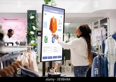 Woman buyer selecting clothes on board, using interactive touch screen monitor in clothing store. Young boutique customer buying fashion items from self ordering kiosk service, retail shop. Stock Photo