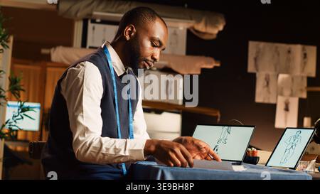 Tailor reviewing designs on tablet, comparing sketches of clothes in atelier workshop. Fashion couturier manufacturing clothing line using tailoring instruments and tools. Handheld shot. Stock Photo