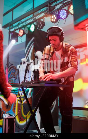 Performer playing techno music at discotheque in nightclub. Young man dj in headphones using digital controller for mixing sound on stage with spotlights at party in club Stock Photo