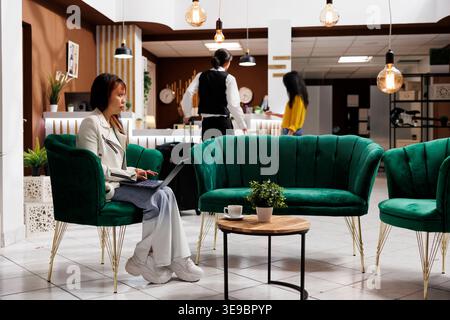 Asian female traveler working on laptop while relaxing in cozy hotel lobby. Young vacationer checks into contemporary resort and unwinds in sitting area before booking room Stock Photo