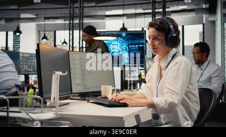 Developer listening music while working in AI startup training predictive models for language processing tasks. Tech expert wearing audio headphones uses technical expertise for IT tasks, camera A Stock Photo
