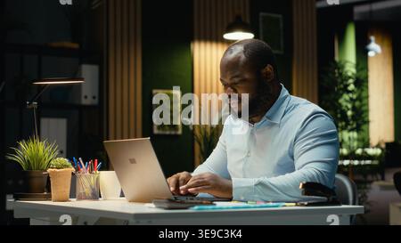 African american guy proofreading a document before transcribing on a textbook, extracting the main ideas from reports on laptop. Expert documenting after hours late at night. Stock Photo