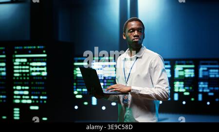 IT developer in server room using laptop to find network issues affecting supercomputer performance and connectivity, leading to slow data transfer rates and poor client communication Stock Photo