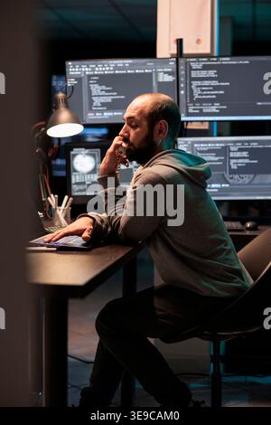 IT engineer answering landline phone call in office, talking about coding html script on cord telephone line. Working late at night to develop new computer system and app servers. Stock Photo