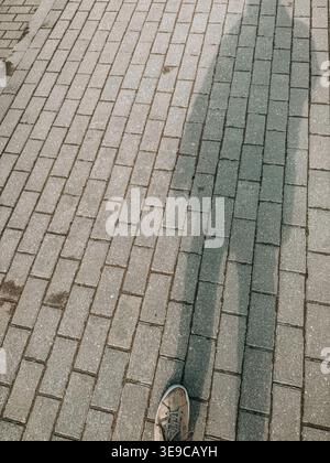 Close-up of a person’s foot stepping on patterned brick pavement with a contrasting dark strip. Minimal urban composition symbolizing direction, choic Stock Photo