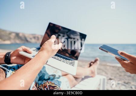 Laptop Beach Work - Man using laptop on the beach while another man uses a smartphone. Stock Photo
