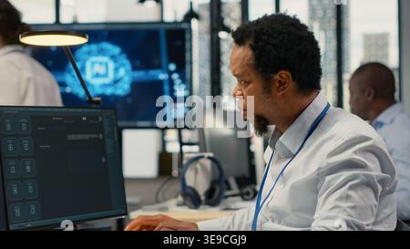 Technician in office solving tasks with artificial intelligence LLM chatbot answering prompts. African american man at work using PC to interact with large language model program, camera B Stock Photo