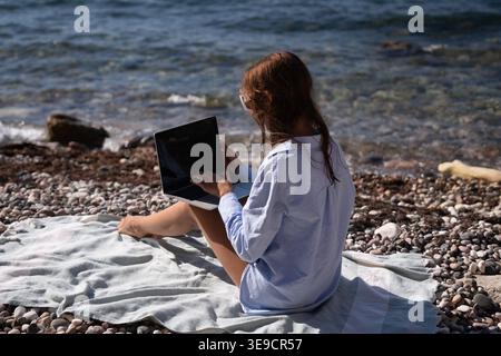 Remote work beach laptop. Woman sits by the sea typing on a computer enjoying sunny day. Stock Photo