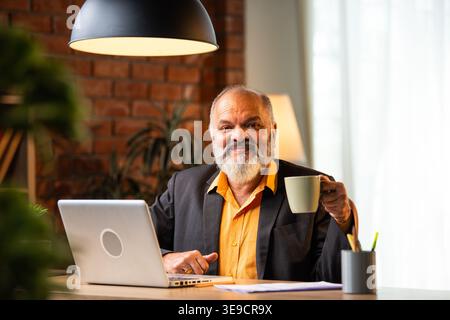 Indian senior businessman or corporate executive standing at his desk, drinking coffee or tea from a mug while working on a laptop in a cozy office en Stock Photo