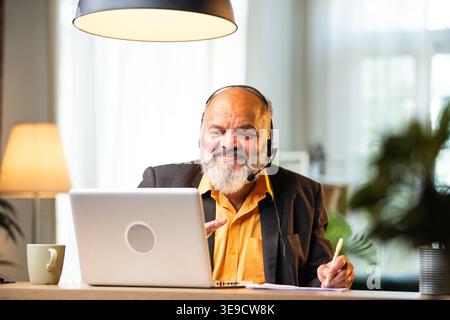 Indian senior businessman or executive wearing a headset with a microphone, taking handwritten notes while actively working on his laptop at a desk in Stock Photo