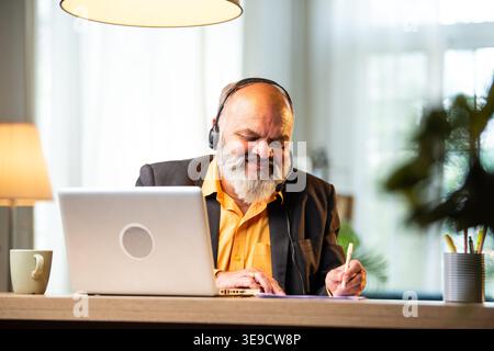 Indian senior businessman or executive wearing a headset with a microphone, taking handwritten notes while actively working on his laptop at a desk in Stock Photo