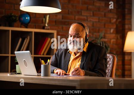 Indian senior businessman or executive wearing a headset with a microphone, taking handwritten notes while actively working on his laptop at a desk in Stock Photo