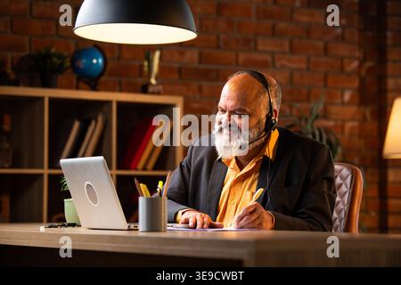Indian senior businessman or executive wearing a headset with a microphone, taking handwritten notes while actively working on his laptop at a desk in Stock Photo