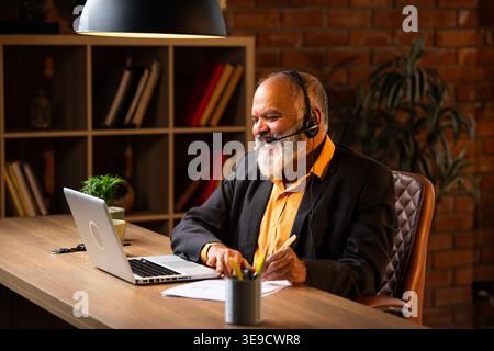 Indian senior businessman or executive wearing a headset with a microphone, taking handwritten notes while actively working on his laptop at a desk in Stock Photo
