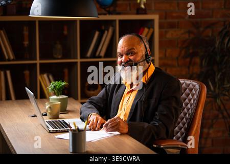 Indian senior businessman or executive wearing a headset with a microphone, taking handwritten notes while actively working on his laptop at a desk in Stock Photo
