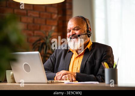 Smiling Indian senior businessman or corporate executive wearing a headset with a microphone, taking handwritten notes while working on his laptop at Stock Photo