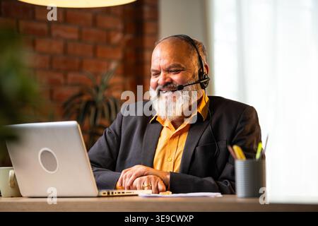 Smiling Indian senior businessman or corporate executive wearing a headset with a microphone, taking handwritten notes while working on his laptop at Stock Photo