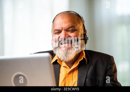 Smiling Indian senior businessman or corporate executive wearing a headset with a microphone, taking handwritten notes while working on his laptop at Stock Photo