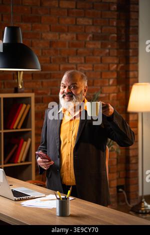 Indian senior businessman or corporate executive multitasking by speaking on a smartphone while holding a coffee or tea mug at his desk in a cozy offi Stock Photo