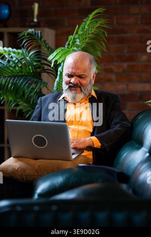 Indian senior businessman or corporate executive working on his laptop while sitting comfortably on a sofa in a cozy office environment with professio Stock Photo