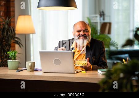 Indian senior businessman or corporate executive having a light snack while multitasking on a laptop and participating in a video call at his desk in Stock Photo