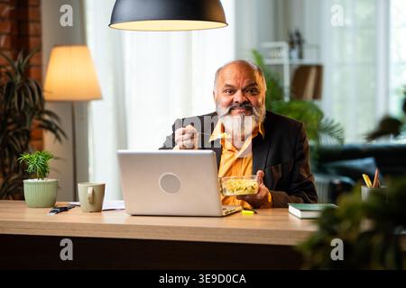 Indian senior businessman or corporate executive having a light snack while multitasking on a laptop and participating in a video call at his desk in Stock Photo
