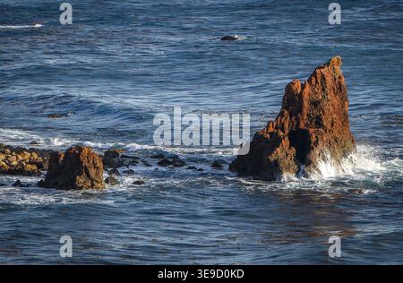 Ocean waves breaking against rugged coastal sea stack with surrounding rocks and deep blue water. Stock Photo