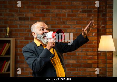 Indian senior businessman or corporate executive happily announcing news or shouting through a megaphone while standing against a red brick wall in a Stock Photo