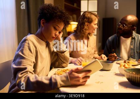 Teenager boy using smartphone during family dinner, ignoring parents and showing screen distraction at home. Ideal for parenting, digital addiction, family communication campaigns Stock Photo