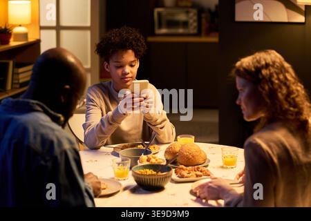 Teenager using smartphone during family dinner while parents sitting at table, showing screen distraction and digital overuse. Useful for parenting, screen time, family lifestyle marketing Stock Photo