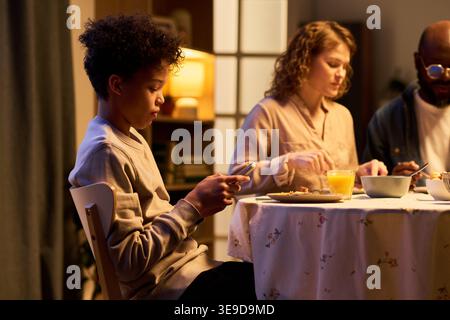 Black teenager using smartphone during family dinner, showing screen distraction and digital dependence. Useful for parenting, technology habits, family communication campaigns Stock Photo
