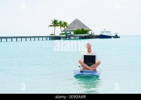 Smiling, successful woman with laptop surfing on a SUP  Success. Flexible remote work, female work-life balance concept. Stock Photo