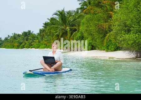 Smiling, successful woman with laptop surfing on a SUP  Success. Flexible remote work, female work-life balance concept. Stock Photo
