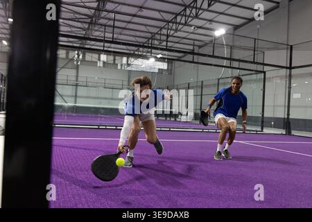 Two men diving, playing padel on purple court with black rackets, yellow ball in sports hall Stock Photo
